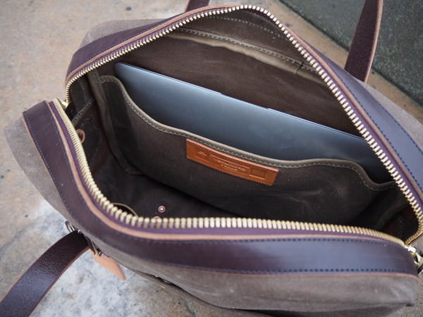 Brass zipper at top of light brown fabric bag is opened.  Dark brown leather trims brass zipper.  Laptop pocket on one side of bag with laptop in it.  Vermilyea Pelle logo sewn to pocket.