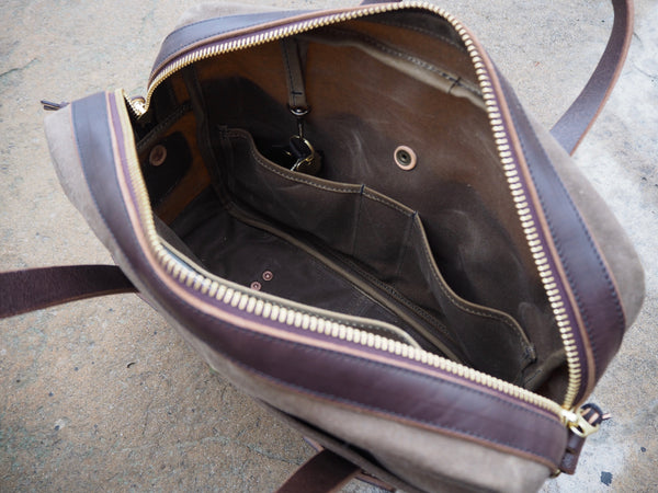 Brass zipper at top of light brown fabric bag is opened.  Dark brown leather trims brass zipper.  Two small pockets on one side of bag.  Antique brass key clip attached inside the top of bag.