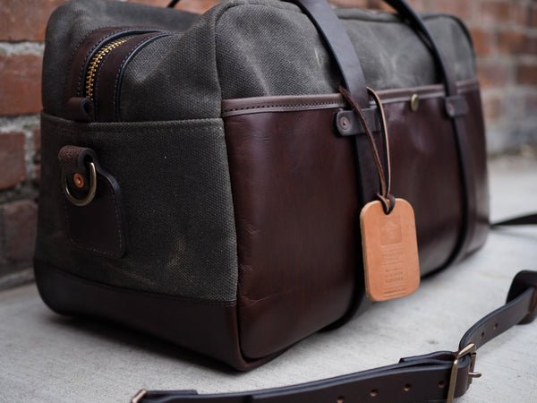 Dark green canvas bag measuring 20 inches long, 10 inches wide and 12 inches tall. Dark brown leather bottom and front pocket. Two dark brown leather straps riveted to front pocket with copper rivets that becomes the handle. Dark brown leather handle wrap. YKK Brass zipper. Dark brown leather shoulder strap with copper rivets and antique brass swivel snaps.