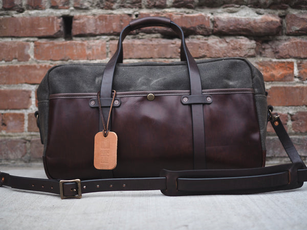 Dark green canvas bag measuring 20 inches long, 10 inches wide and 12 inches tall. Dark brown leather bottom and front pocket. Two dark brown leather straps riveted to front pocket with copper rivets that becomes the handle. Dark brown leather handle wrap. YKK Brass zipper. Dark brown leather shoulder strap with copper rivets and antique brass swivel snaps.