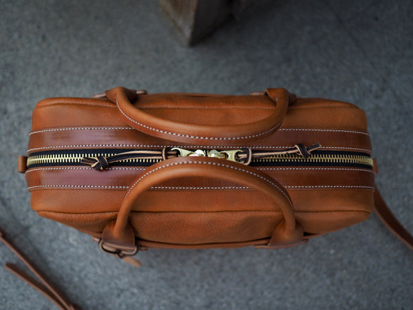 Top of light brown leather briefcase has YKK brass zipper with a light brown leather strap sewn on either side of zipper. Rolled light brown leather handles.
