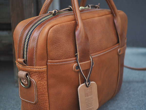 Light brown leather briefcase measuring 15.5 inches long, 11.75 inches tall and 4.5 inches wide. Two light brown leather straps riveted to front leather pocket that turn into rolled handles. Light brown leather shoulder strap with antique brass roller buckles attached to briefcase.  Antique brass D-ring copper riveted to side of bag.