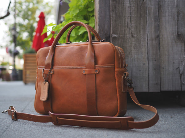 Light brown leather briefcase measuring 15.5 inches long, 11.75 inches tall and 4.5 inches wide. Two light brown leather straps riveted to front leather pocket that turn into rolled handles. Light brown leather shoulder strap with antique brass roller buckles attached to briefcase.