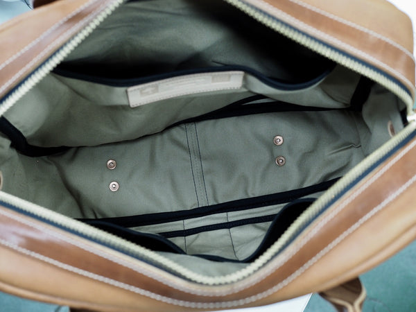 YKK Brass zipper opened of light brown leather briefcase. Inside of bag has 2 pockets on one side and laptop pocket on other side with Vermilyea Pelle logo. 4 copper rivets hammered to bottom of interior. Bag lined in tan canvas duck fabric.